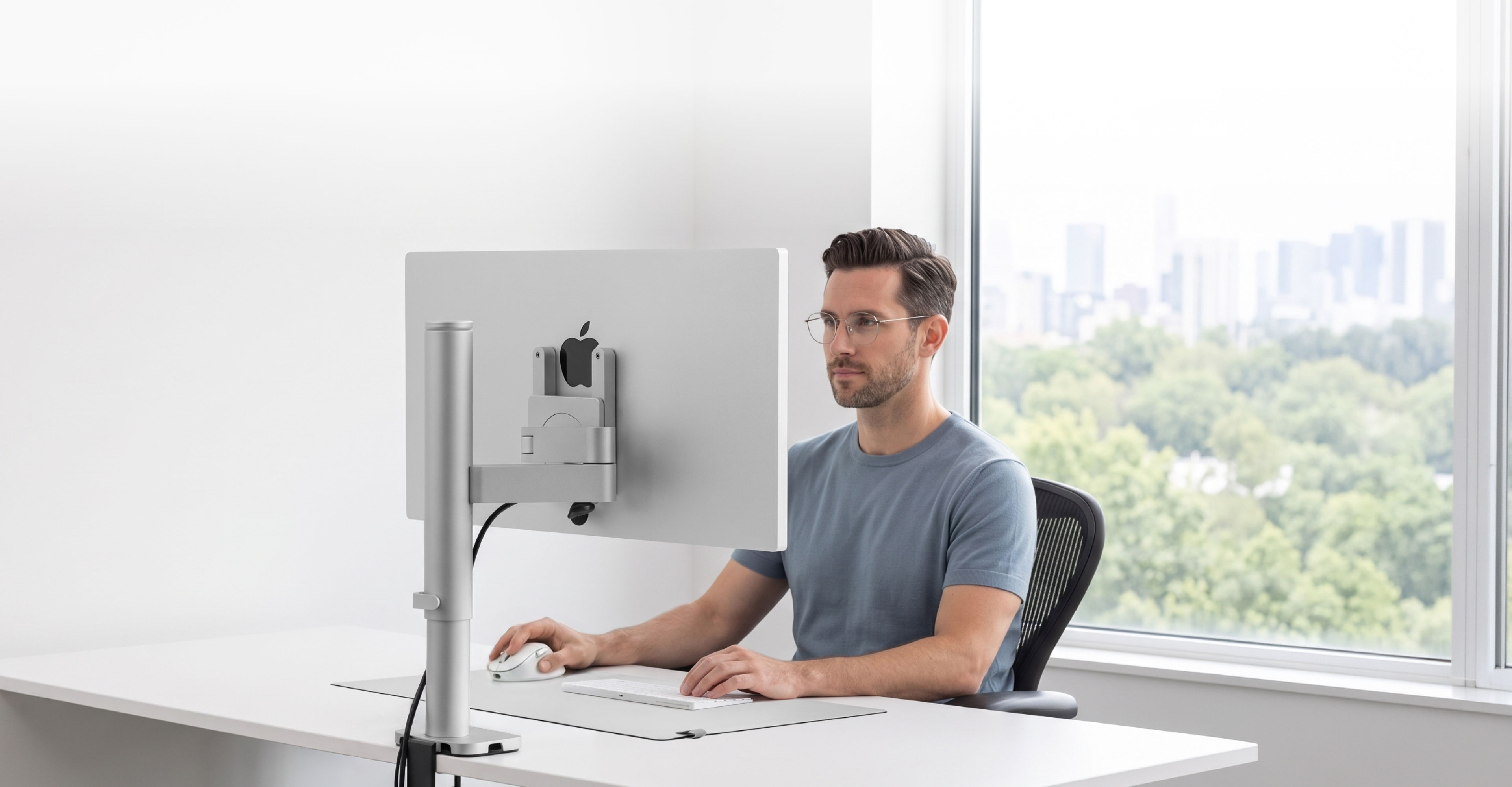 Man working at a bright, minimalist desk setup with Studio Arm supporting an Apple Studio Display, showing a clean and organized workspace with cables hidden through the arm and base. A large window in the background overlooks a green landscape, emphasizing the focused and clutter-free working environment enabled by integrated cable management.