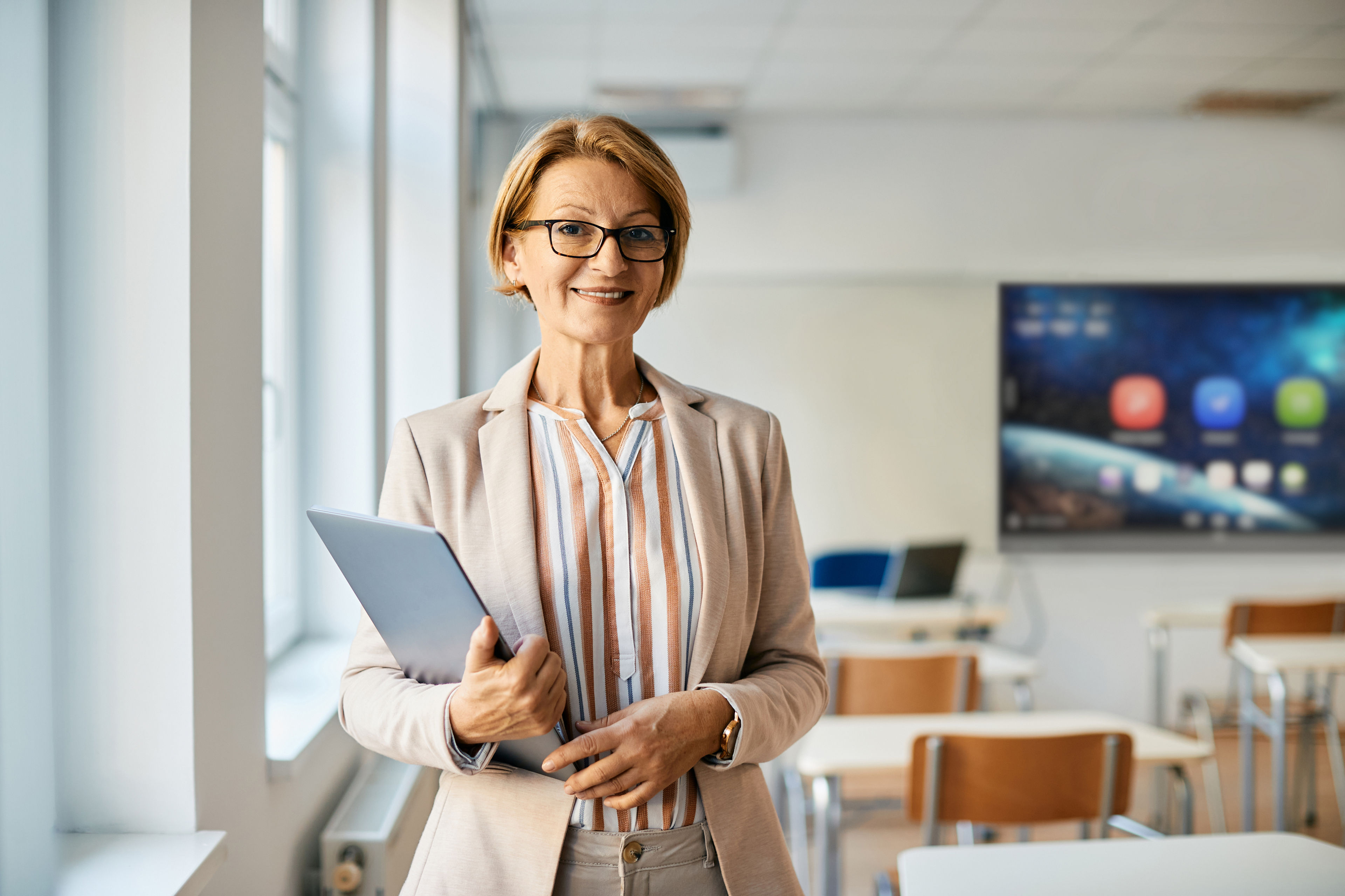 Portrait of smiling mature teacher with laptop in the classroom.