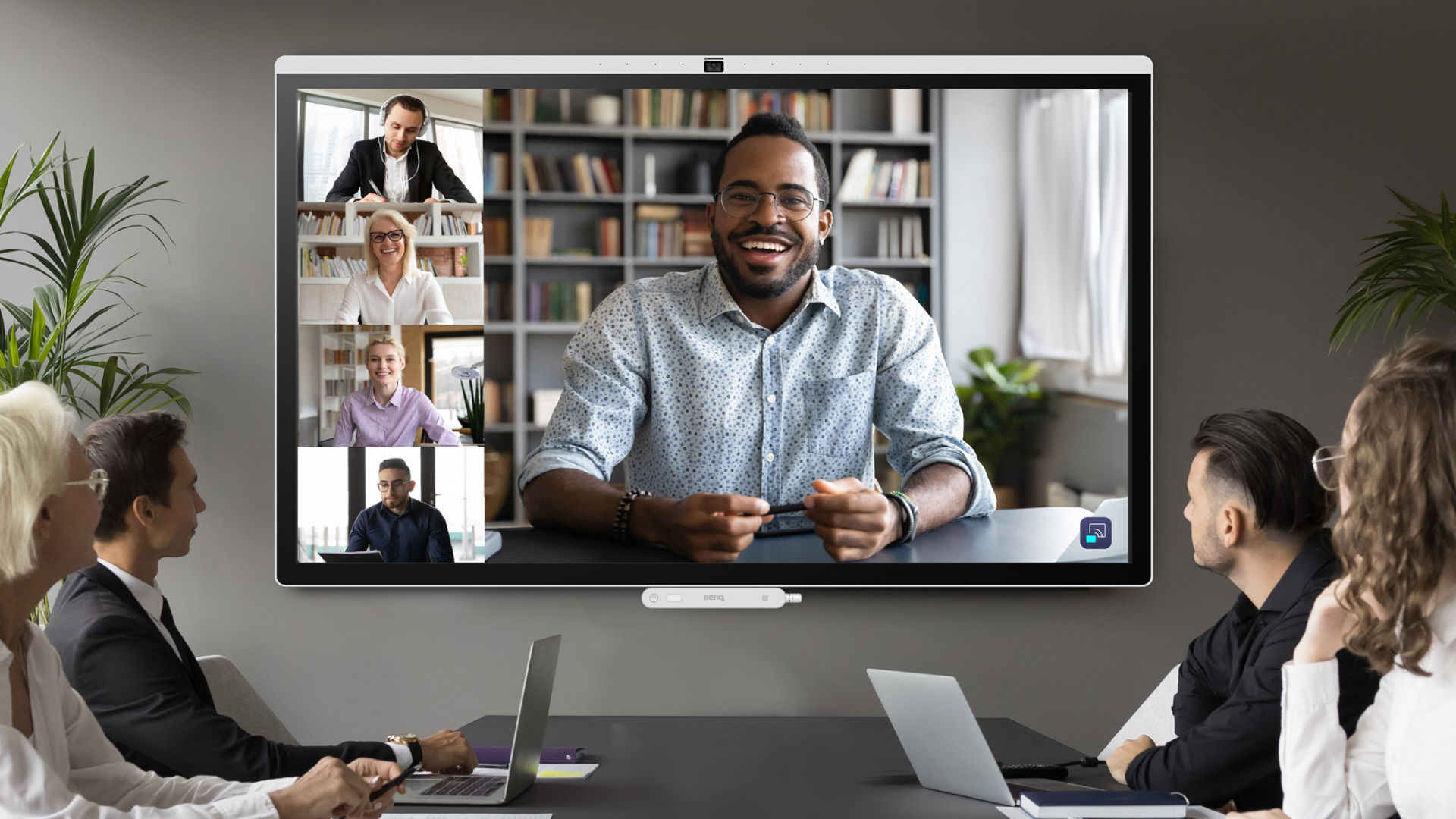 A large BenQ display in a corporate boardroom showing a multi-person video conference call with clear, high-resolution visuals to support remote team meetings.