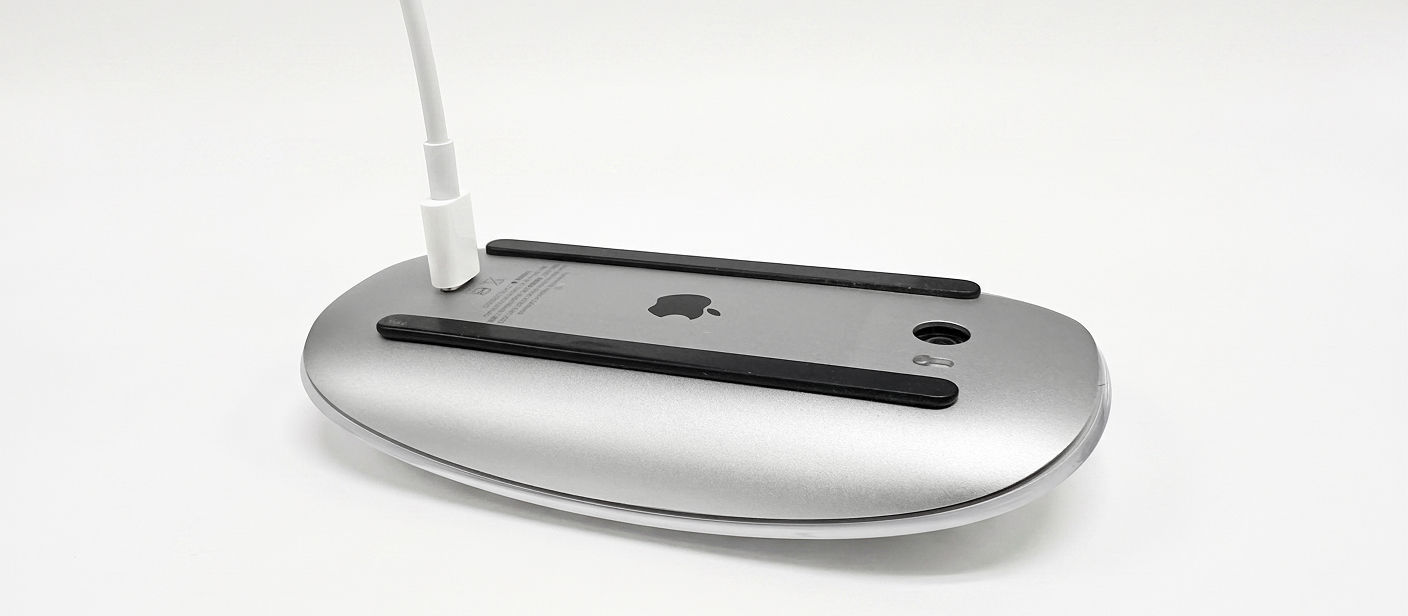 Magic Mouse shown charging upside down through its bottom-mounted port, making it unavailable for use while plugged in.