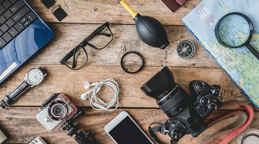 There are various objects and electronic products on the table such as laptop, digital camera, SLR camera, mobile phone, earphone, glasses, compass and reading glass.