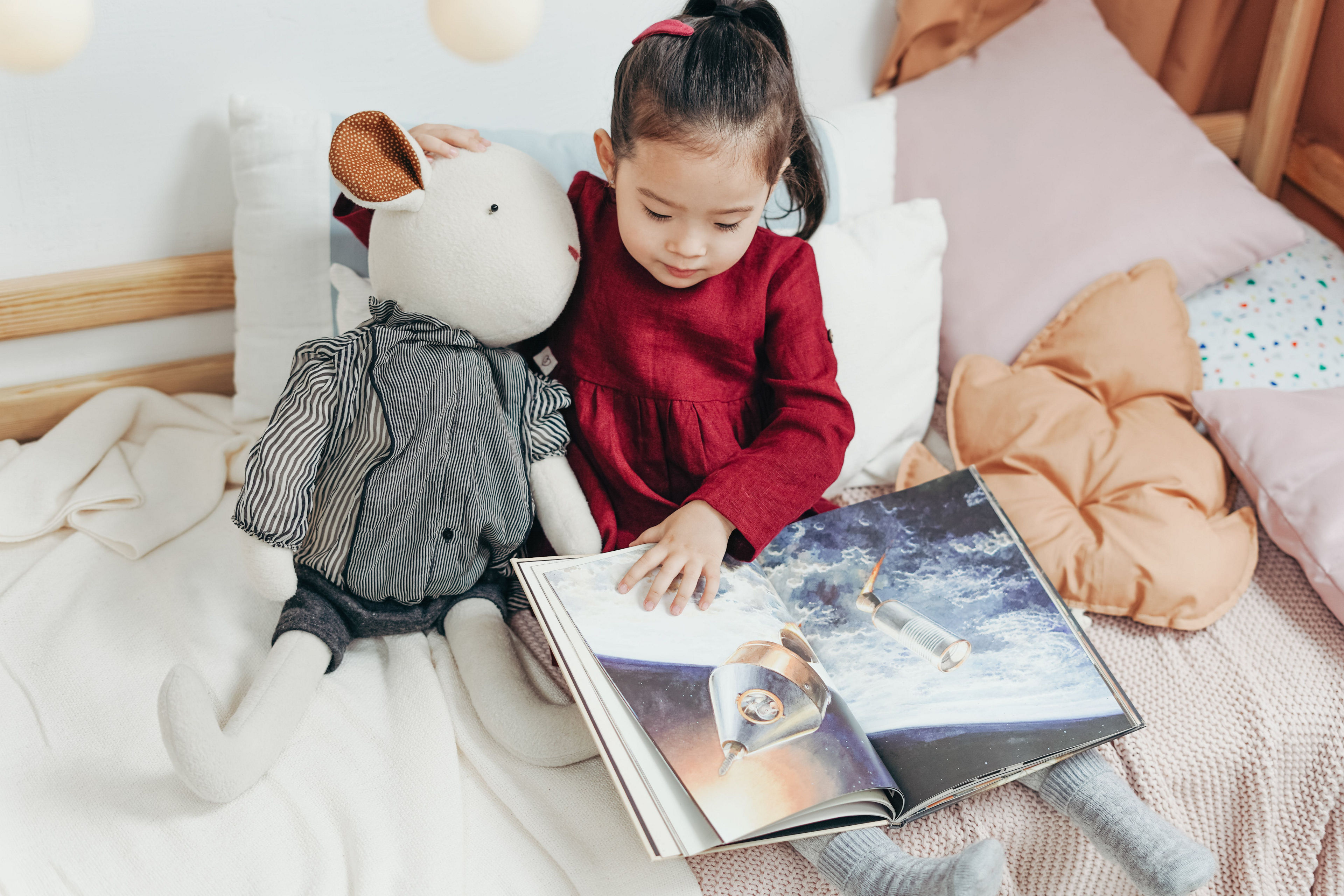 A little girl wearing a red dress reading a book on bed