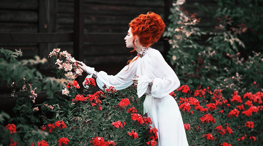 A woman with orange hair and white dress is touching flowers in the garden.