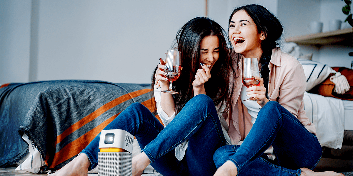 There are two girls enjoying big screen entertainment on a mini portable projector GV1 in a small room.